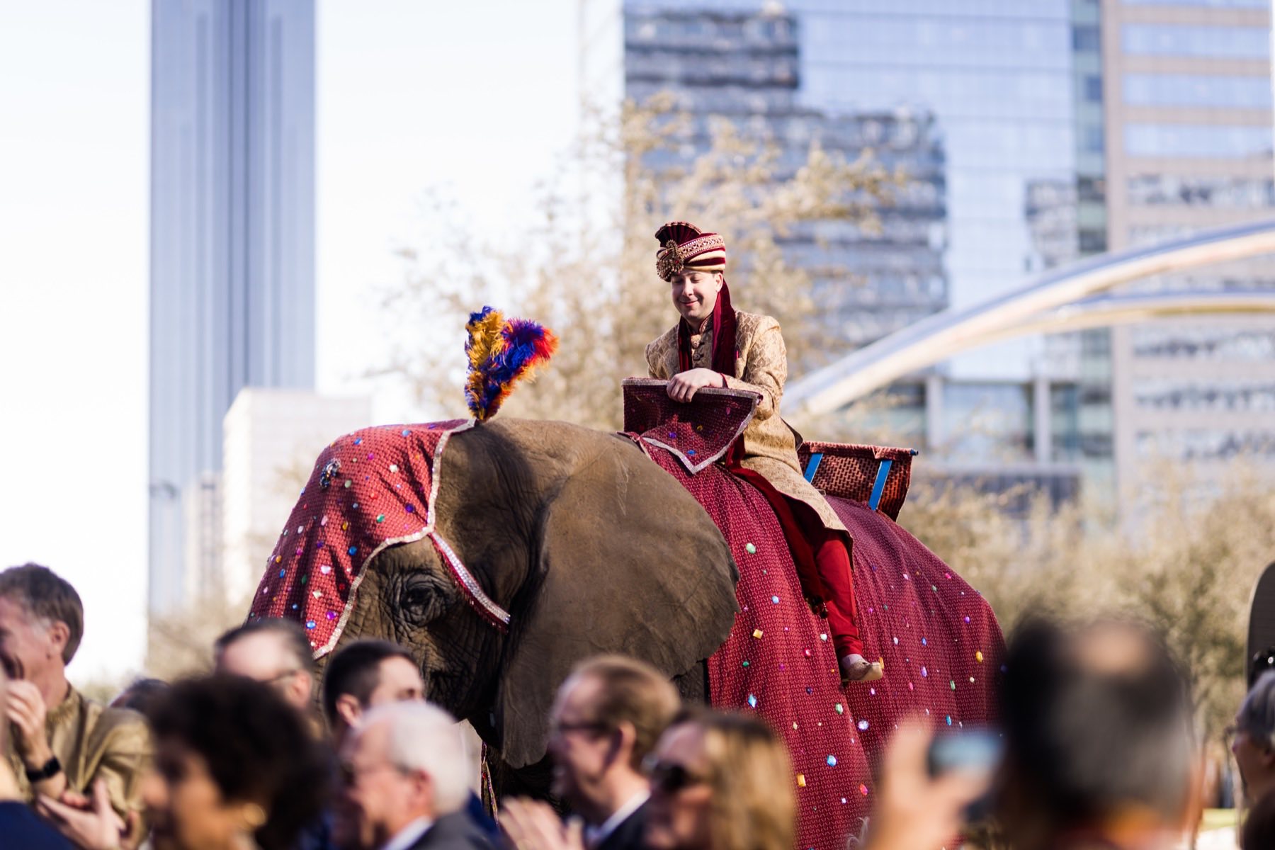 Groom in gold sherwani arriving on a decorated elephant against city skyline backdrop