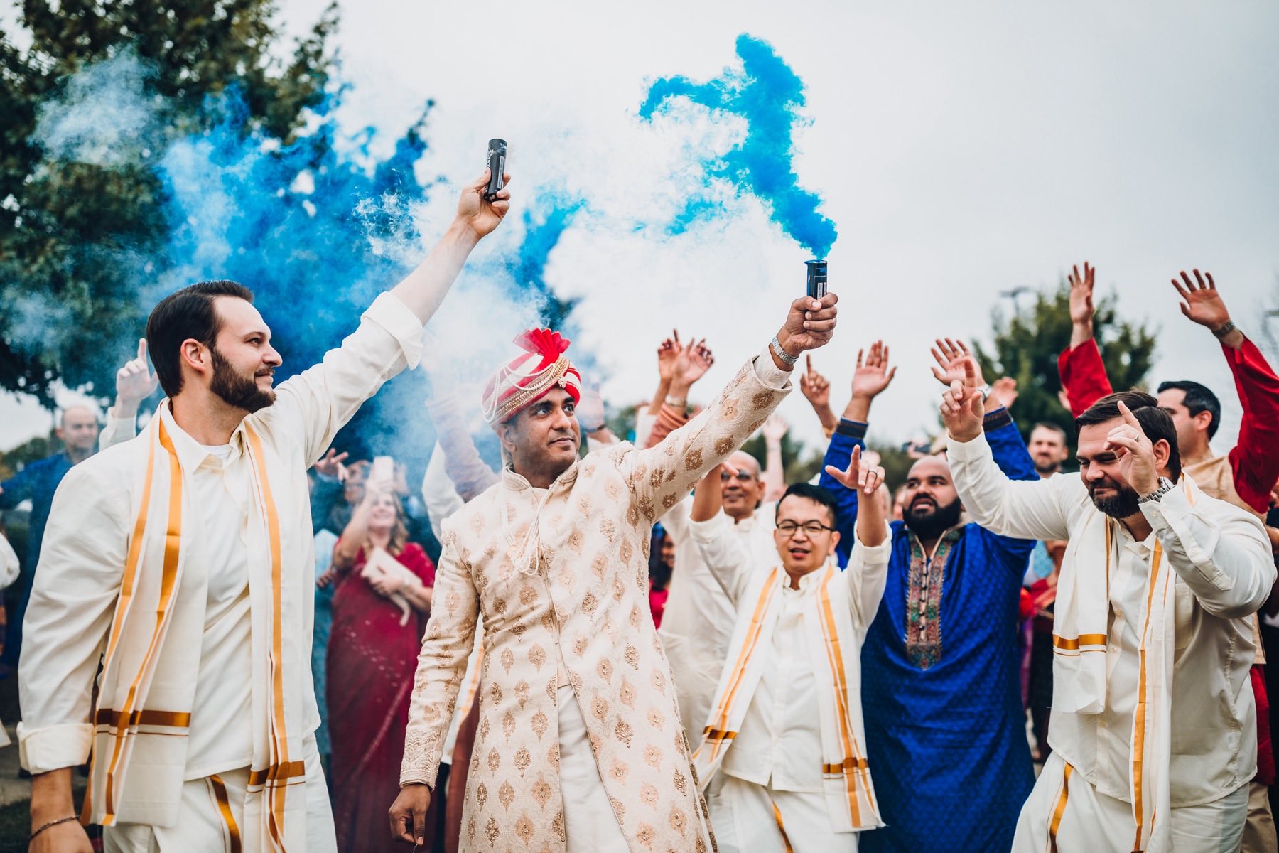 Groom holding blue smoke bomb aloft surrounded by cheering groomsmen at baraat