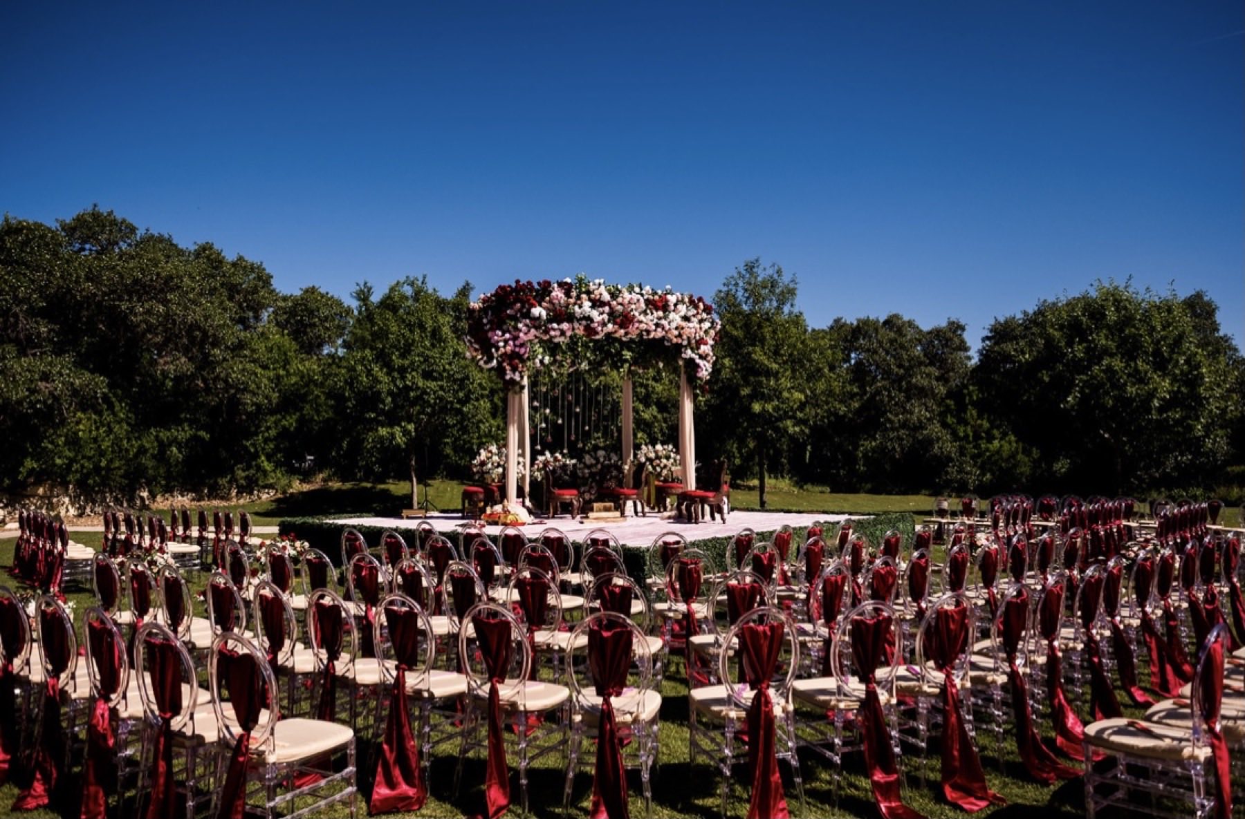 Outdoor ceremony under blue sky with lush floral mandap and red-sashed ghost chairs