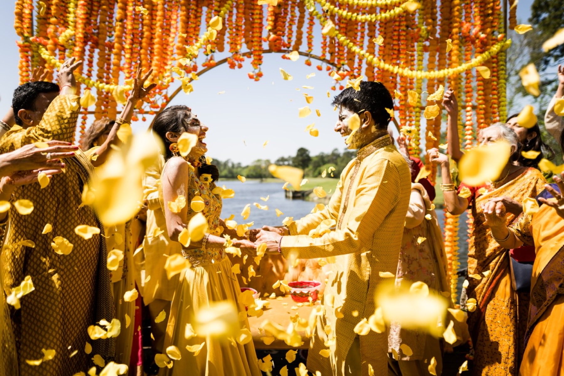 Couple holding hands as yellow flower petals are tossed at haldi ceremony by the waterside
