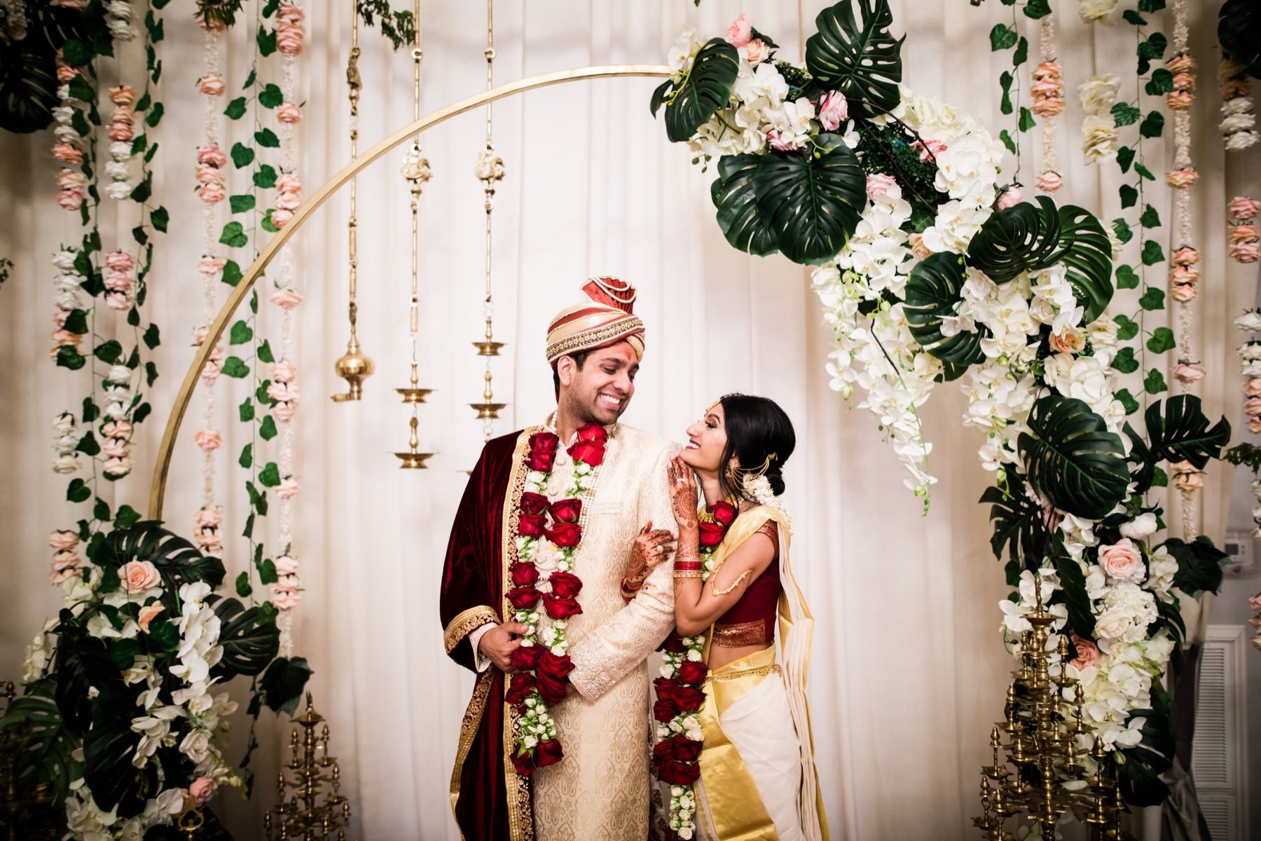 Couple smiling at each other beneath tropical monstera and rose floral arch
