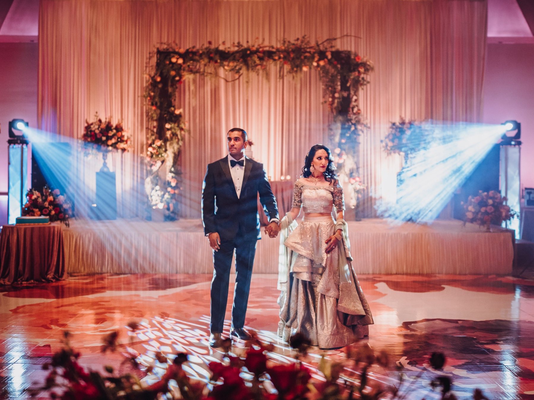 Couple holding hands on spotlit dance floor with warm floral stage backdrop