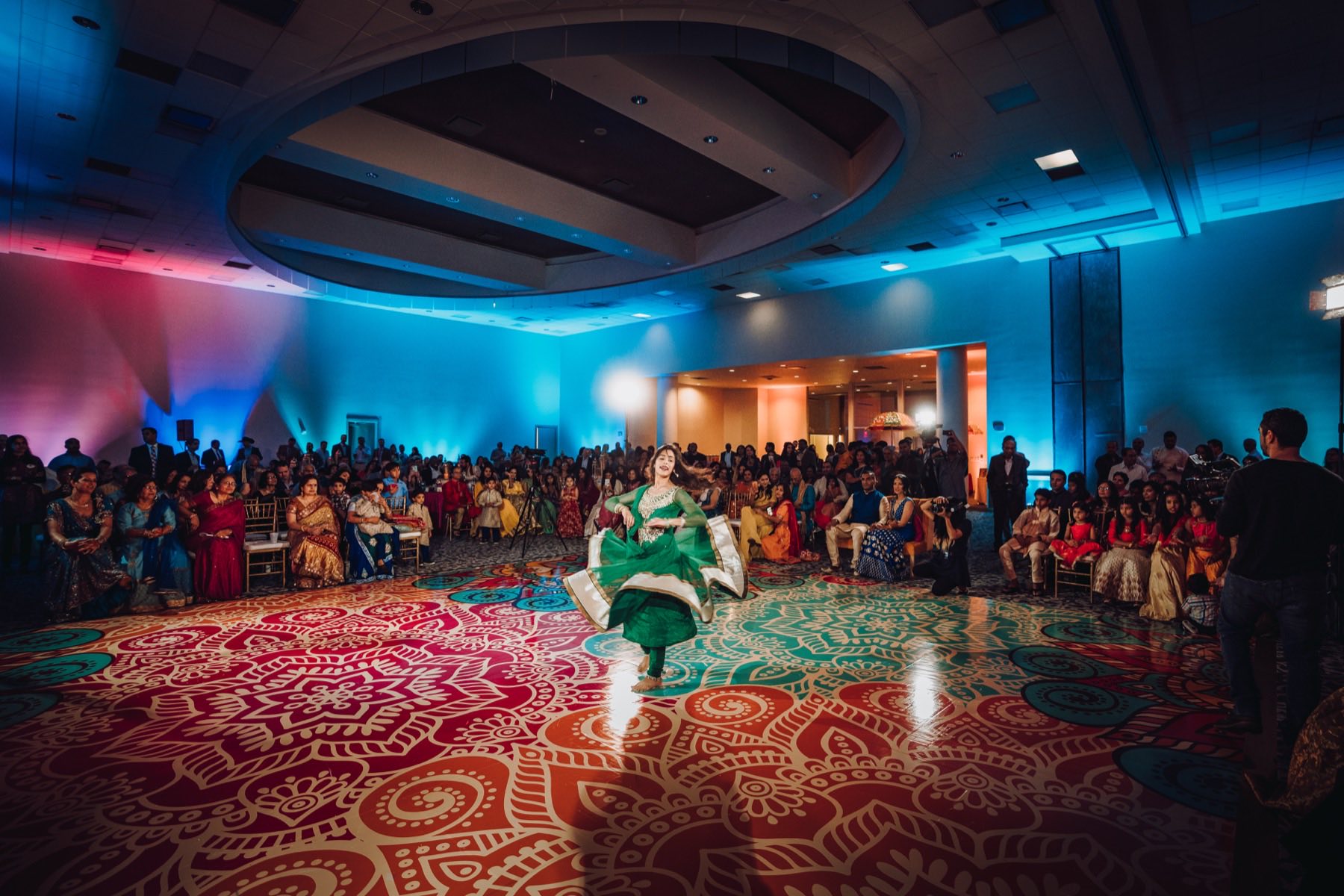 Dancer spinning on ornate carpet at sangeet under blue and pink uplighting in full ballroom