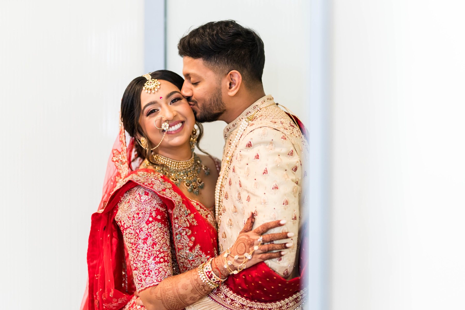 Groom kissing bride on the cheek as she smiles in red lehenga and gold jewelry